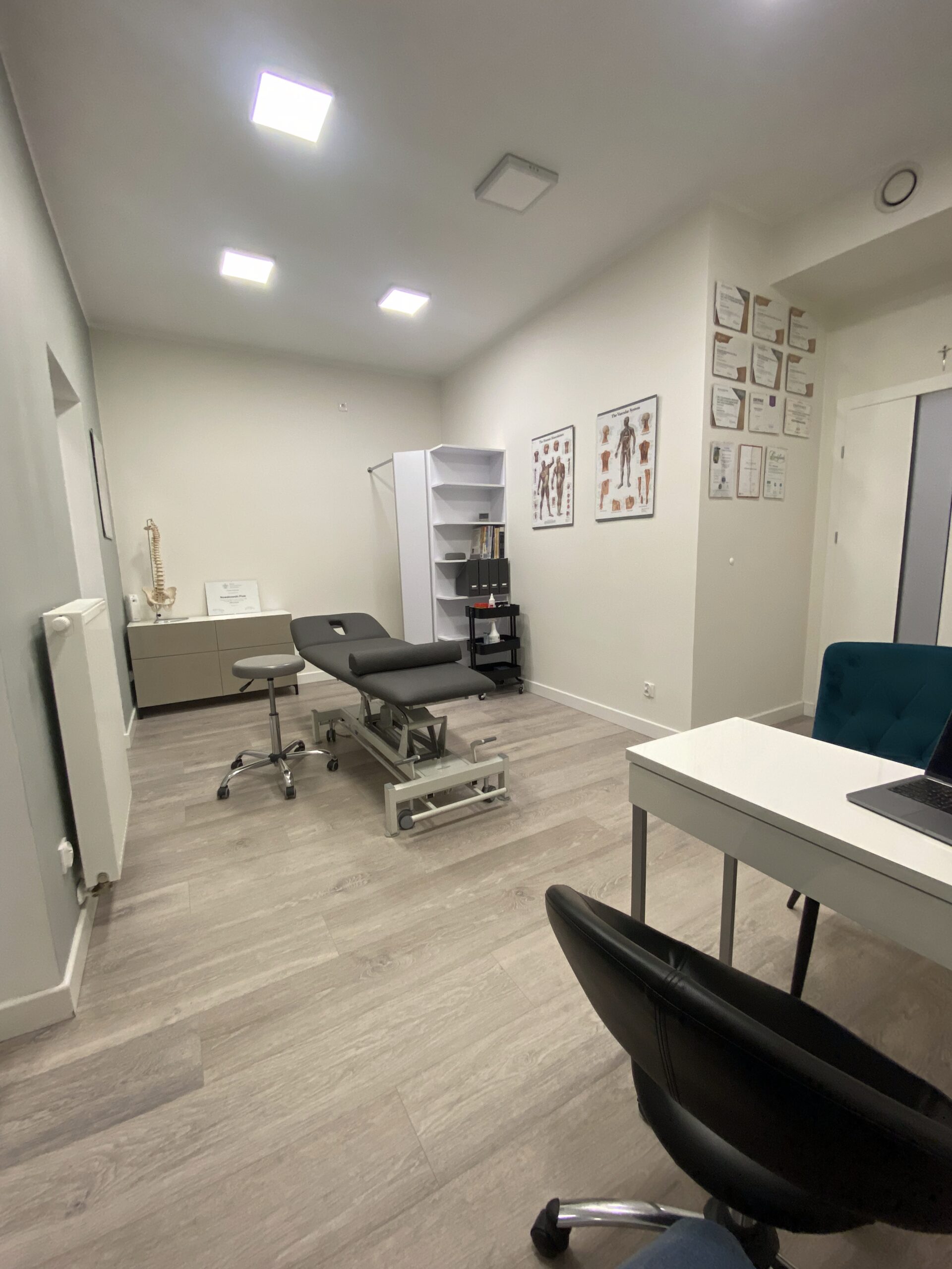 Jasny gabinet fizjoterapeutyczny z leżanką, taboretami, biurkiem i materiałami edukacyjnymi na ścianach.