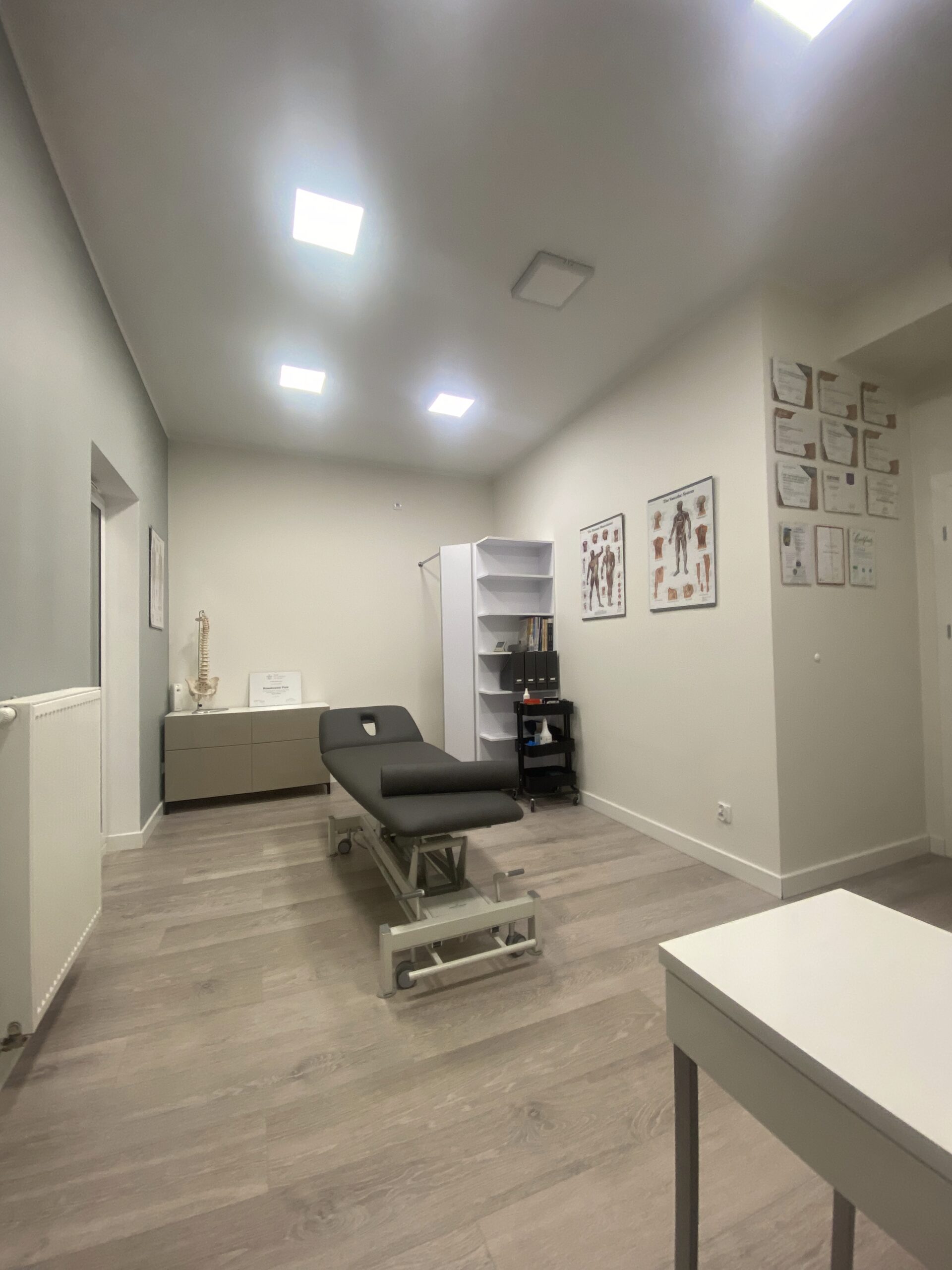 Jasny gabinet fizjoterapeutyczny z leżanką, taboretami, biurkiem i materiałami edukacyjnymi na ścianach