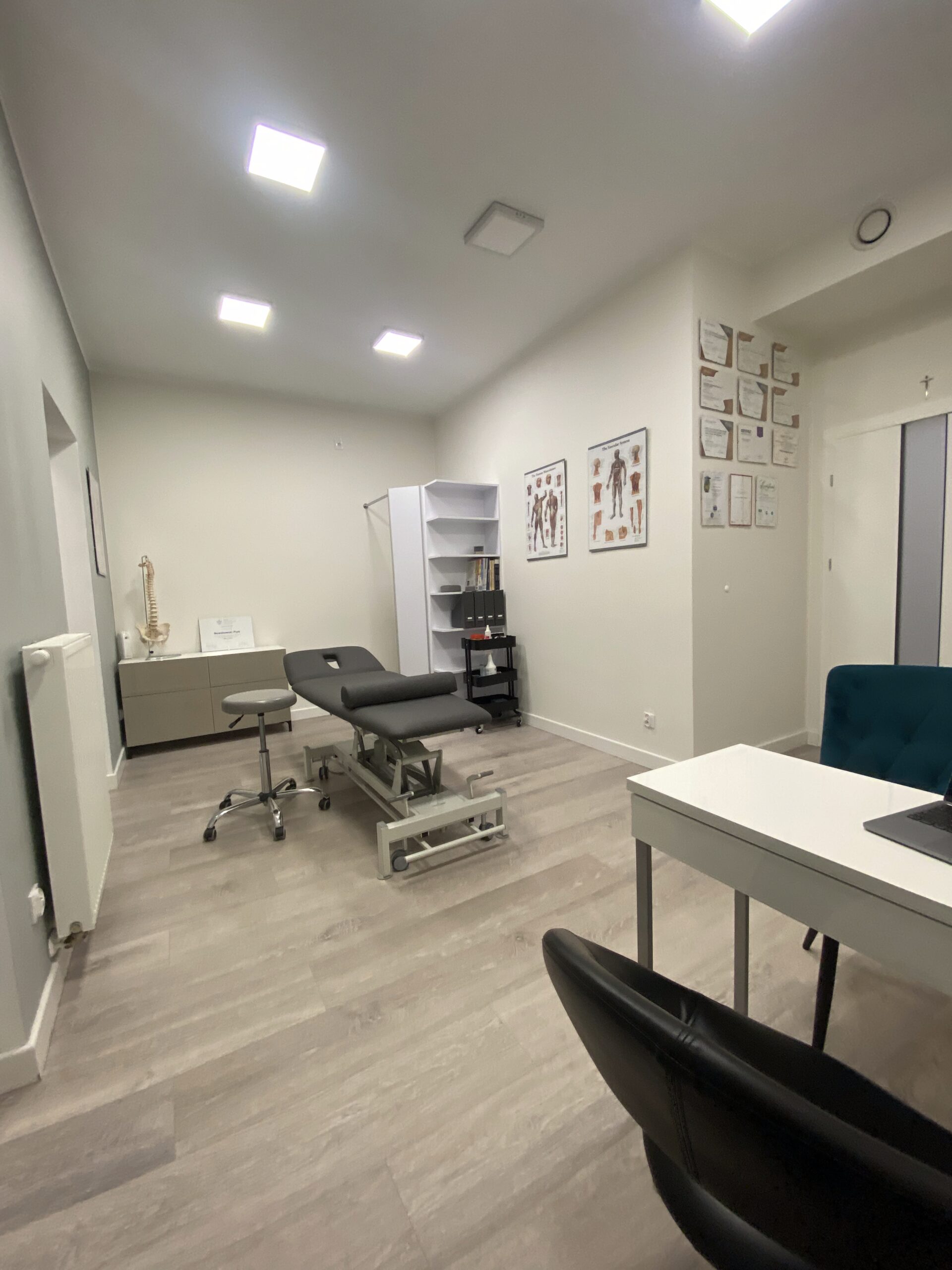 Jasny gabinet fizjoterapeutyczny z leżanką, taboretami, biurkiem i materiałami edukacyjnymi na ścianach.